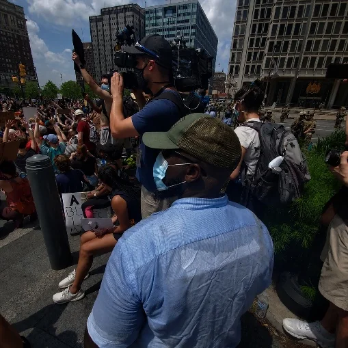 A protective professional positioned beside a camera operator in a crowded public environment.