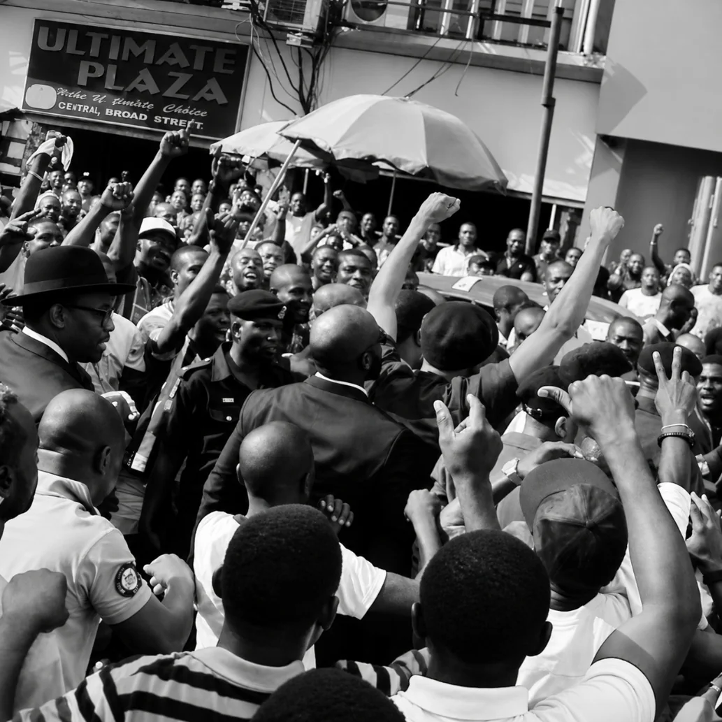 A protection detail moving through a dense public crowd around a visible principal.